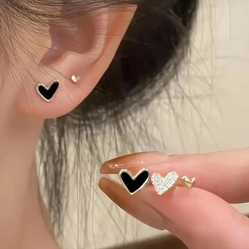 Three pairs of heart-shaped alloy earrings in gold, black, black and white, and pink colours on a white background.
