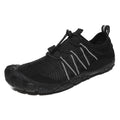 Unisex outdoor water shoes in black with mesh upper and rubber sole, shown on rocky riverbank.