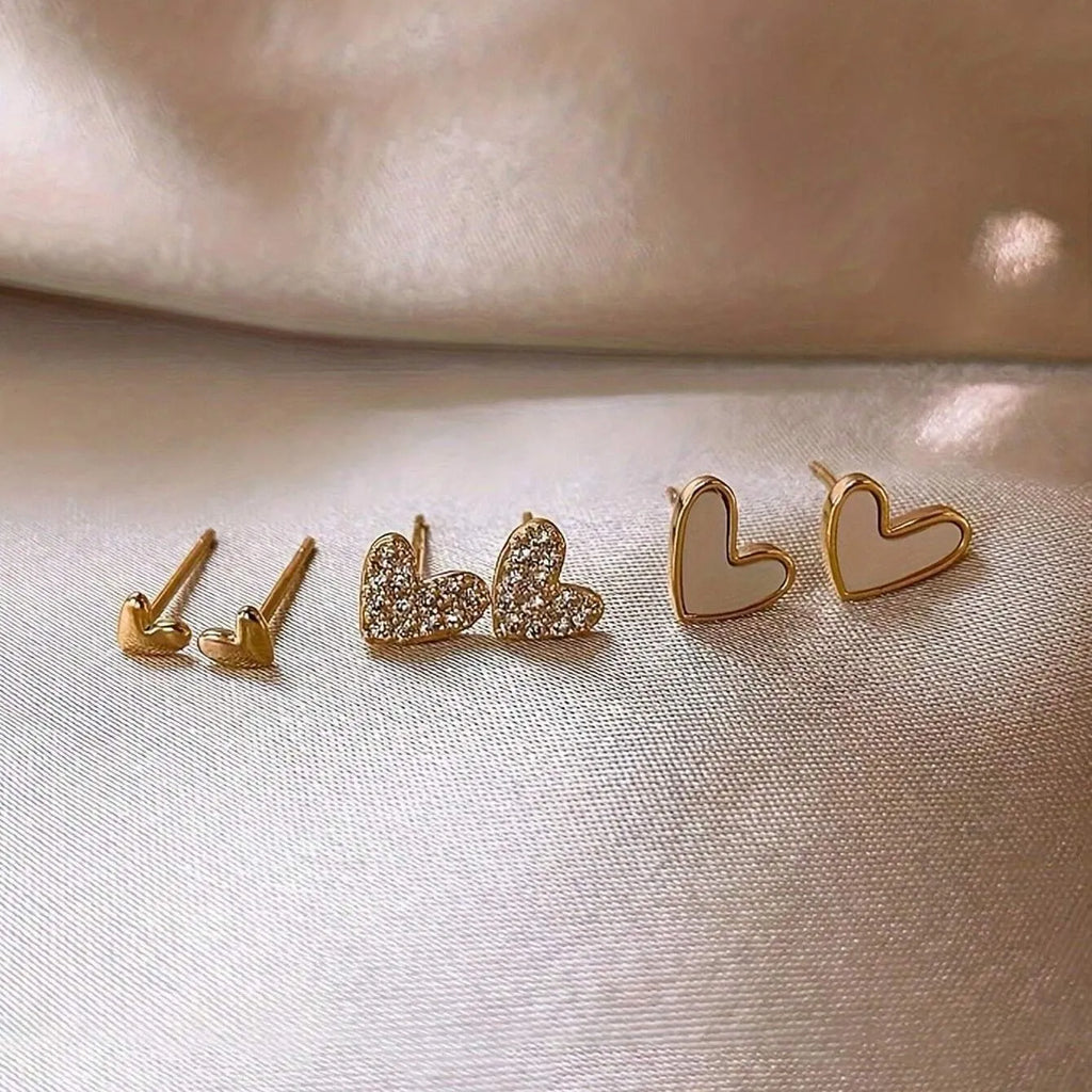 Three pairs of heart-shaped alloy earrings in gold, black, black and white, and pink colours on a white background.
