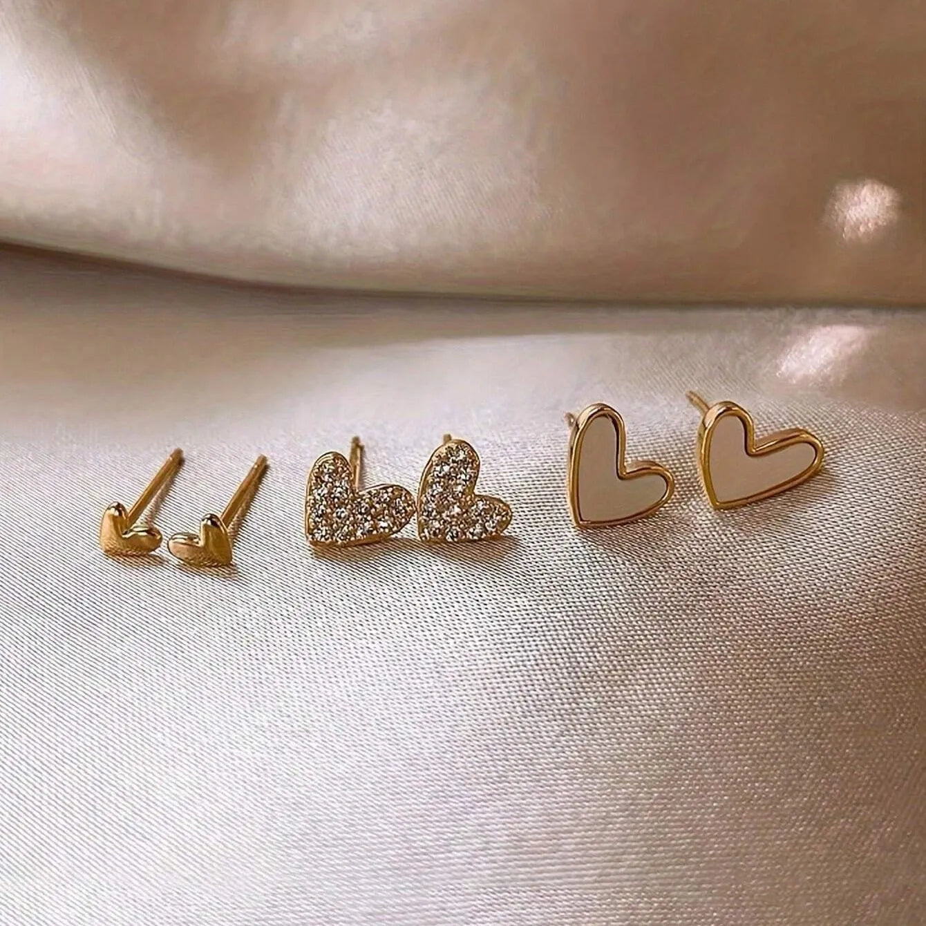 Three pairs of heart-shaped alloy earrings in gold, black, black and white, and pink colours on a white background.