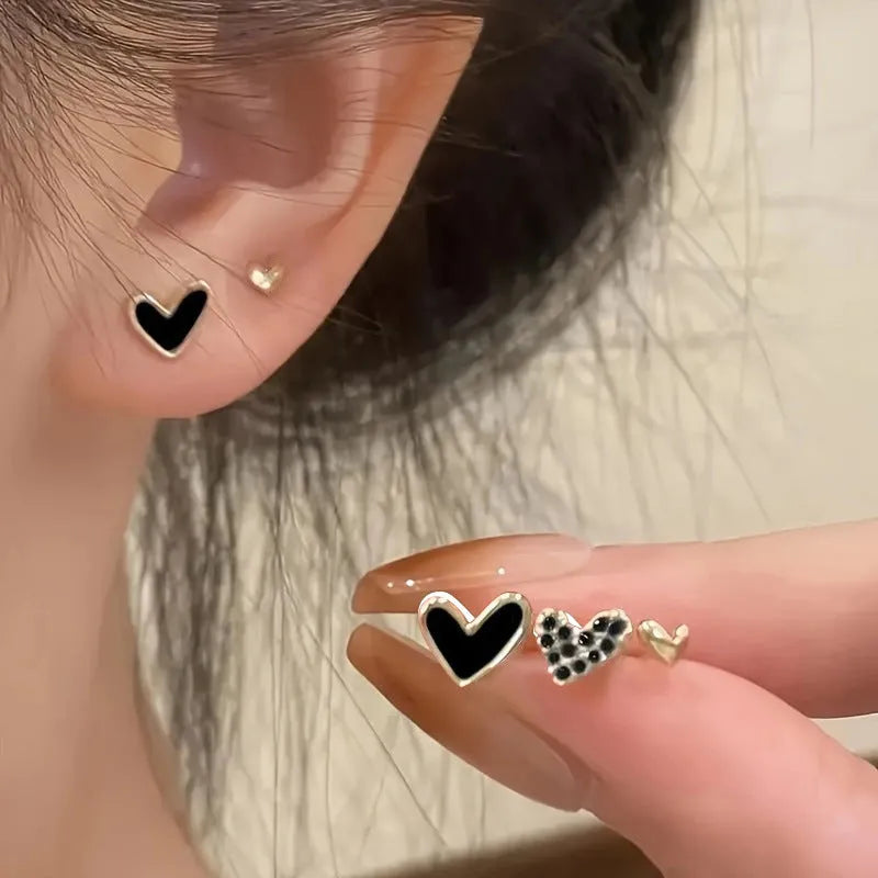 Three pairs of heart-shaped alloy earrings in gold, black, black and white, and pink colours on a white background.